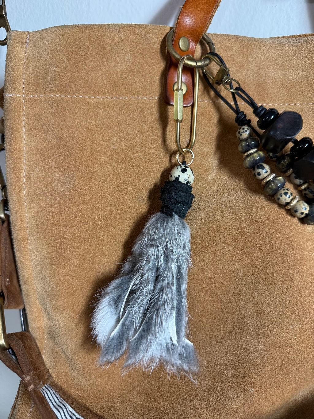 A close-up of a tan suede or leather bag with a hanging fur tassel keychain. The tassel includes a gray and white fur, a black leather wrapped section, a small dalmatian Jasper bead, and a brass carabiner.