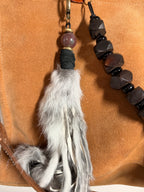 A close-up of a beaded tassel accessory with a white and gray faux-fur tassel, a round brown bead, black wrapped cord, and dark faceted beads on a tan suede-like background.