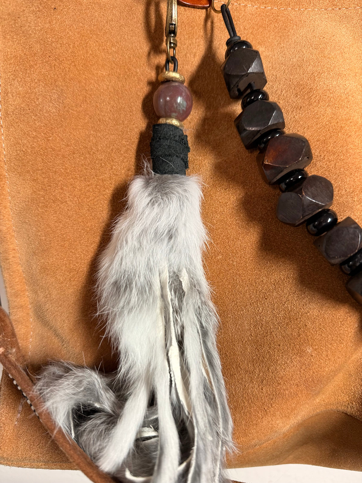 A close-up of a beaded tassel accessory with a white and gray faux-fur tassel, a round brown bead, black wrapped cord, and dark faceted beads on a tan suede-like background.