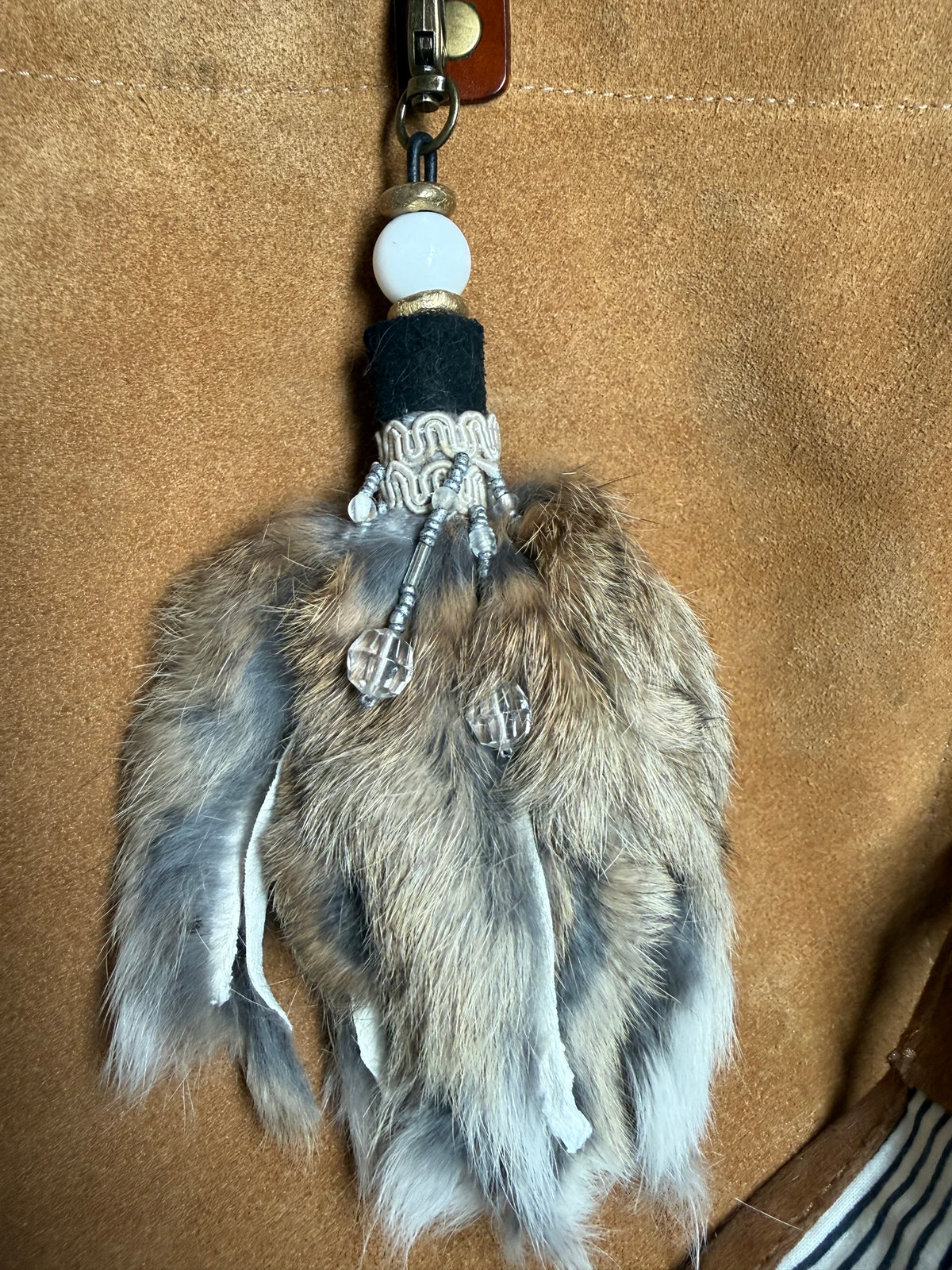 A handmade keychain or bag charm featuring natural brown and gray fur, a black fabric loop, a white braided cord, a white bead, and silver-toned metal accents, shown against a tan suede background.