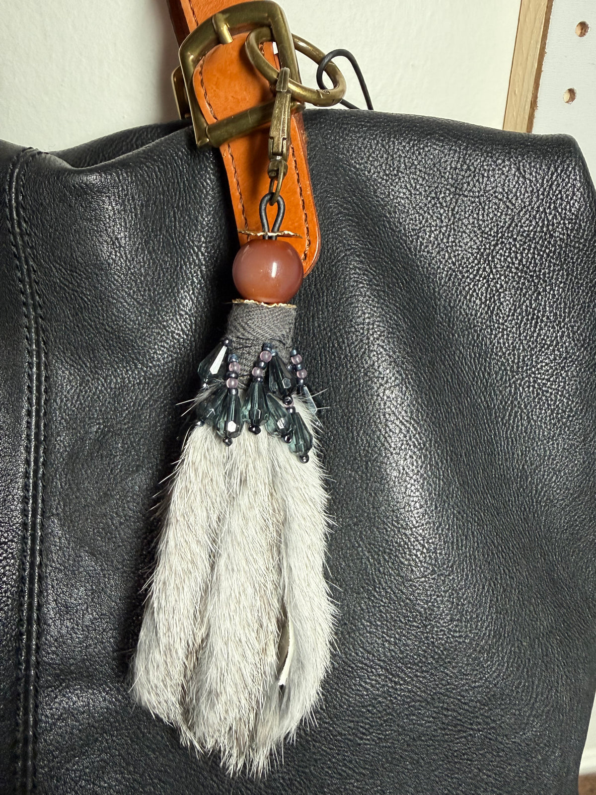 A close-up of a black textured leather bag featuring a decorative tassel charm. The charm includes a light gray fur tassel, a brown stone bead, and a brass buckle attached to an orange-brown leather strap.