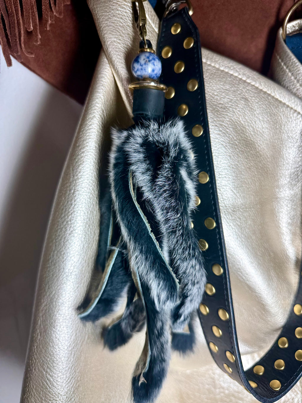 A close-up of a handbag strap with a decorative tassel charm. The charm includes a black leather strap with gold studs, a blue and white speckled bead, and a fluffy black-and-white faux fur tassel.