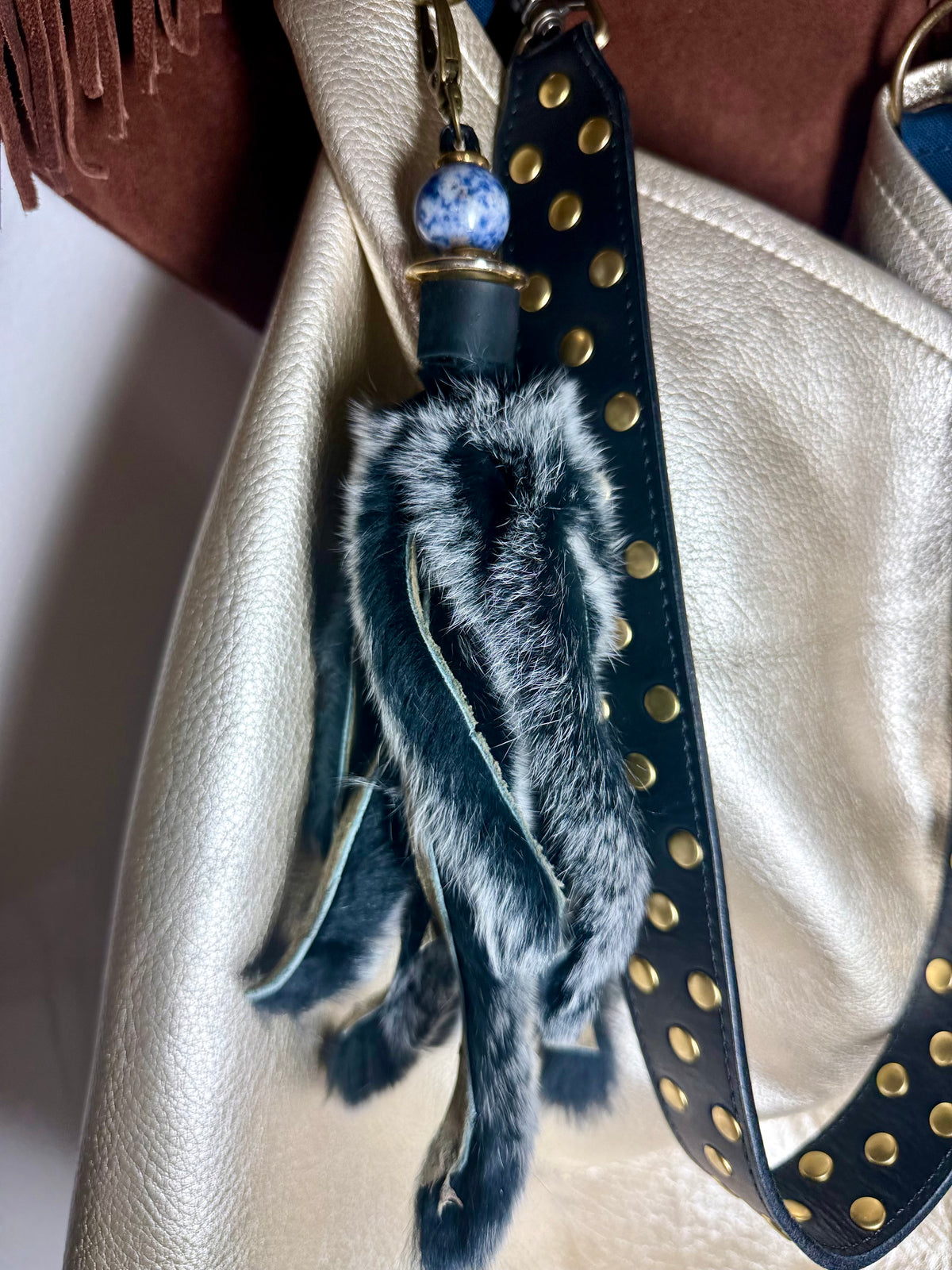 A close-up of a handbag strap with a decorative tassel charm. The charm includes a black leather strap with gold studs, a blue and white speckled bead, and a fluffy black-and-white faux fur tassel.