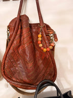 A brown leather hobo shoulder bag with quilted chevron stitching, featuring a beaded wooden keychain charm, displayed against a light background.