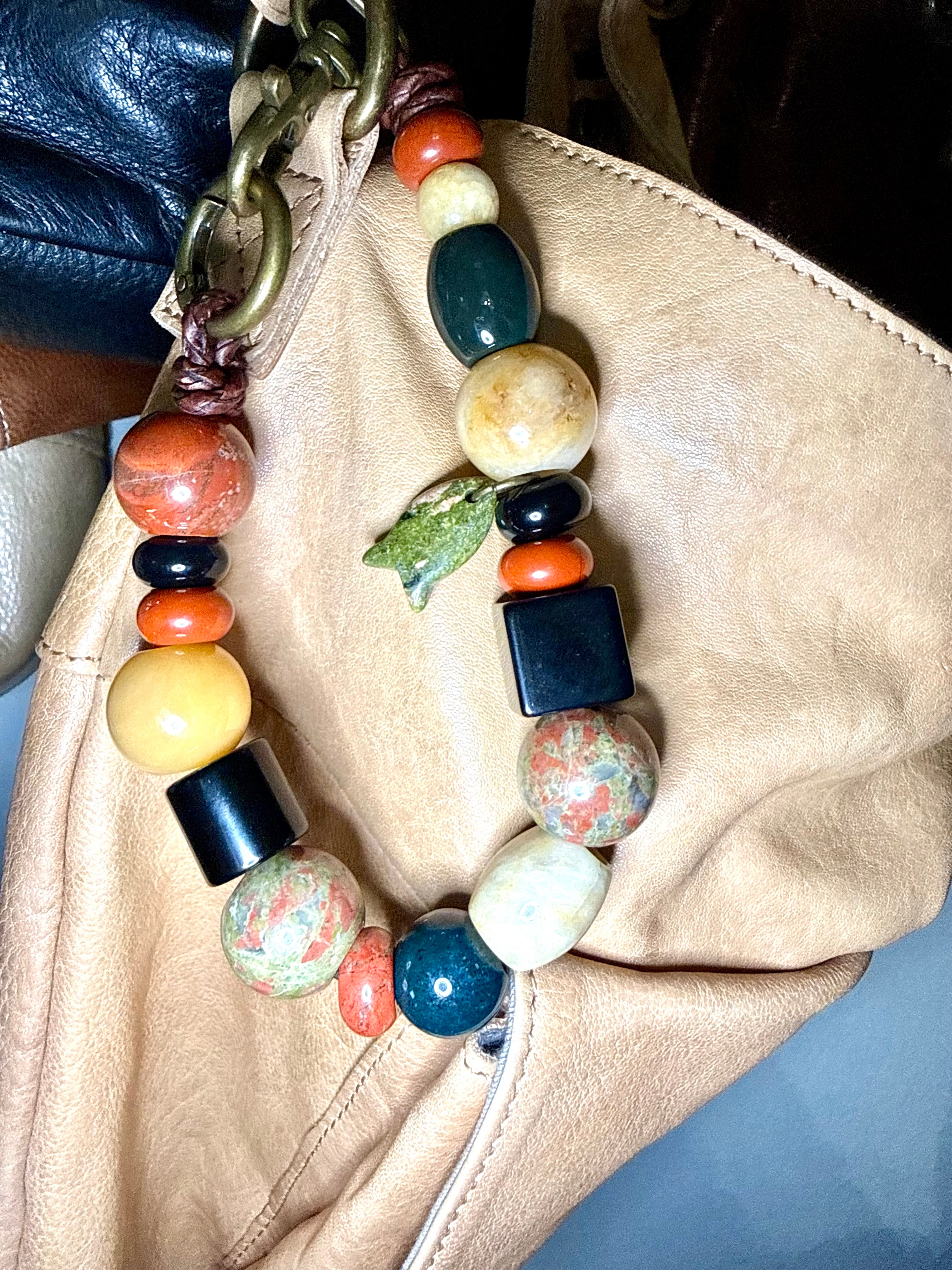 A close-up of a chunky beaded shoulder strap draped over a beige leather bag. The strap features a mix of polished stone beads in orange, yellow, blue, green, black, and white, along with a small green frog charm, attached to a dark blue leather strap with a gold-tone chain.