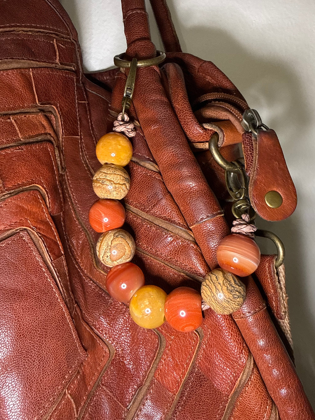 A close-up of a brown leather handbag featuring a decorative beaded strap with orange, yellow, and brown marbled beads attached to brass hardware.