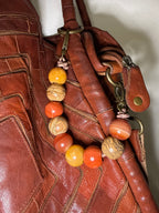 A close-up of a brown leather handbag featuring a decorative beaded strap with orange, yellow, and brown marbled beads attached to brass hardware.