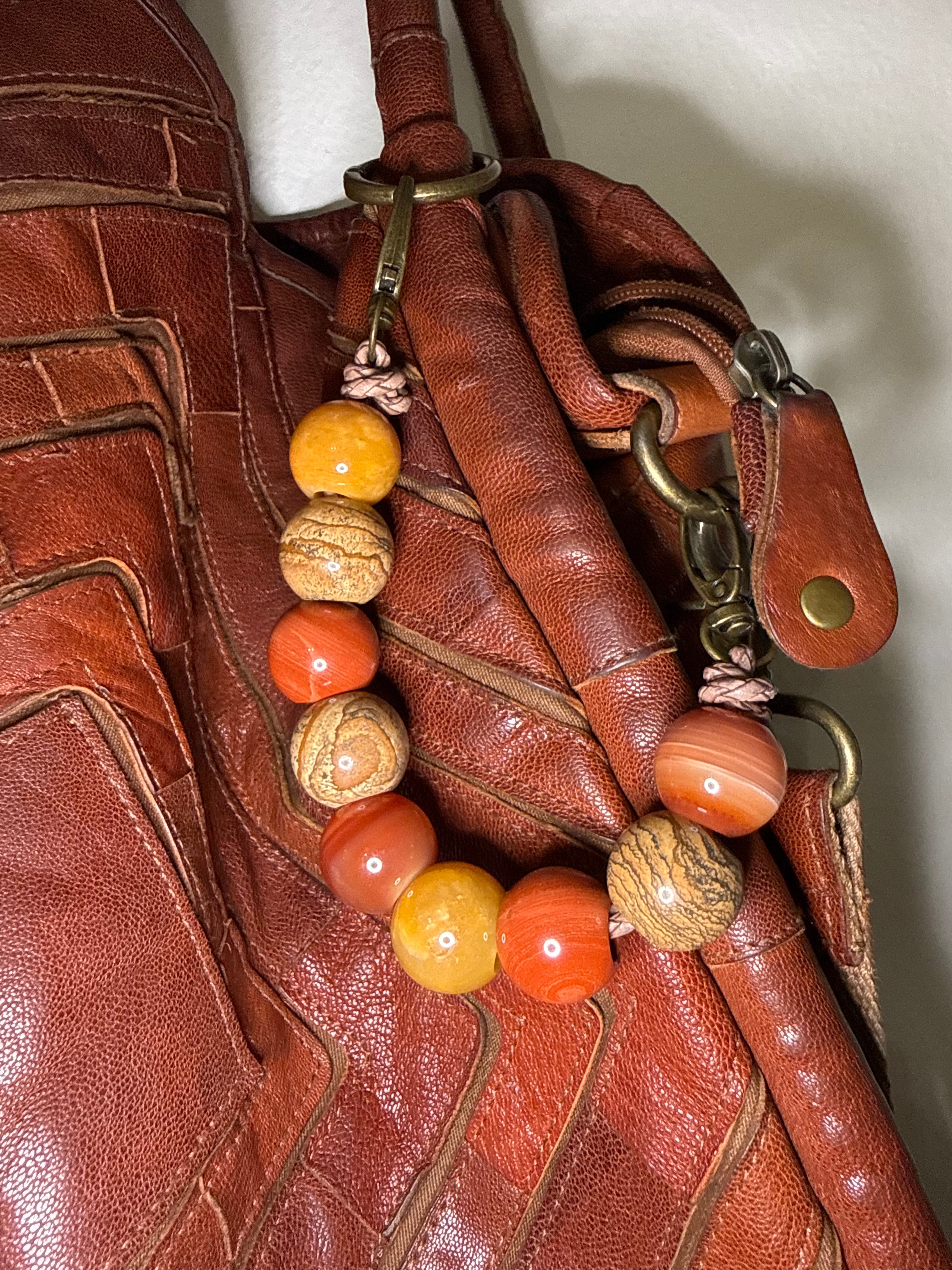 A close-up of a brown leather handbag featuring a decorative beaded strap with orange, yellow, and brown marbled beads attached to brass hardware.