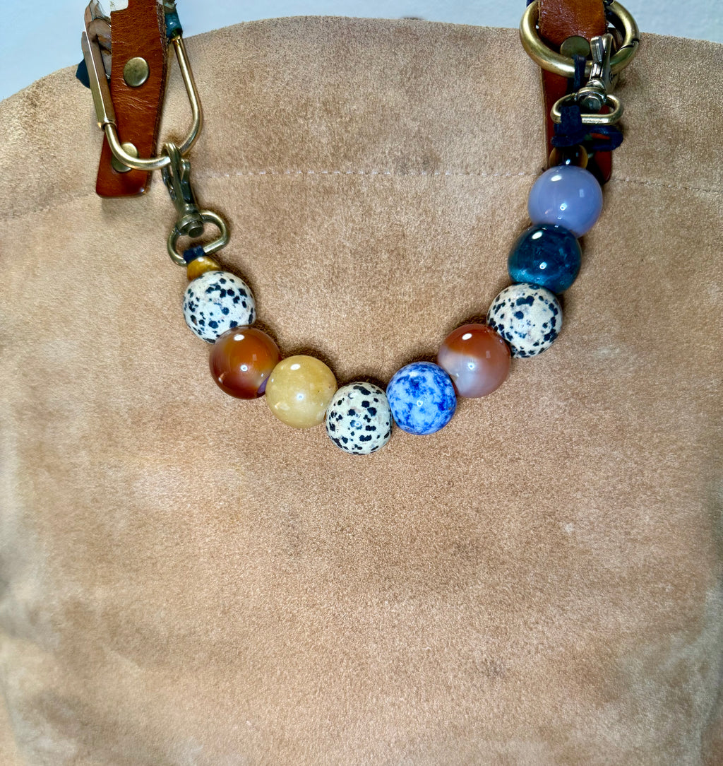A beaded collar with a mix of colorful round beads including blue, white with black spots, brown, and yellow, attached to brown leather straps with brass hardware, displayed on a beige suede-like surface.