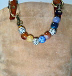 A beaded collar with a mix of colorful round beads including blue, white with black spots, brown, and yellow, attached to brown leather straps with brass hardware, displayed on a beige suede-like surface.
