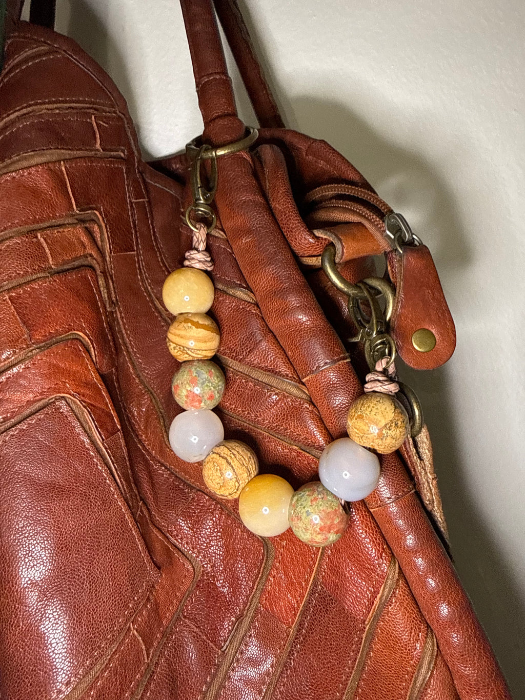 A close-up of a brown leather handbag with a decorative beaded strap featuring natural stone beads in yellow, green, and white, attached with brass hardware.