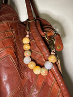 A close-up of a brown leather handbag with a decorative beaded strap featuring natural stone beads in yellow, green, and white, attached with brass hardware.